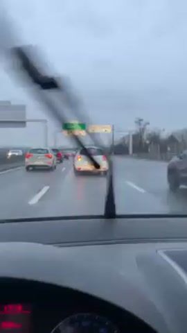 The video shows a car's dashboard view of a wet highway on an overcast day, with traffic moving slowly. The focus is on the vehicles ahead, including a white car and a dark SUV, as they navigate the slick road. Overhead signs provide directions, although the specific destinations are not clearly readable. The car's instrument panel displays the time and a distance reading, suggesting a journey in progress.