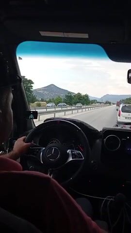 The video is filmed from the interior of a Mercedes-Benz van, looking out through the windshield. A person is driving, their hands visible on the steering wheel. The van is traveling on a highway with other vehicles in front and in the distance. The scenery outside consists of rolling hills, trees, and a cloudy sky. The dashboard of the van, including the infotainment system and a digital clock displaying 1:58, is also visible. In the rearview mirror, reflections show two passengers in the vehicle. A small, colorful air freshener is hanging from the mirror.