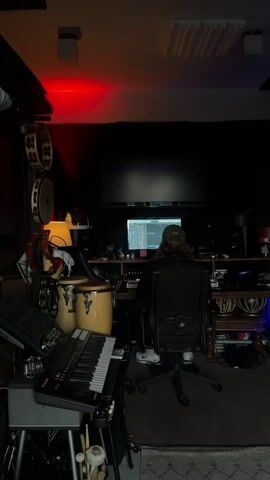 A person sits in a chair facing a computer screen in a dimly lit room that appears to be a recording studio. There are musical instruments visible, including conga drums, a keyboard, and tambourines. Red lighting is present in the upper left corner of the room. The person is wearing a baseball cap and is looking at the screen. The video consists of a single, static shot.