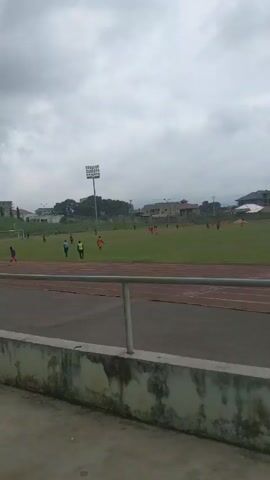 The video shows a soccer field surrounded by a track and seating. There are players on the field wearing different uniforms, suggesting a game in progress. Spectators are sitting in the stands, watching the match. The weather appears overcast, and there are buildings visible in the background. A person walks up the stairs past the seats.
