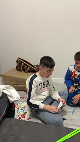 A young boy is sitting on the floor. He is wearing a white sweatshirt with a dark blue stripe on the sleeve and the word "USA" printed on the front and blue jeans. He has short dark hair that is closely cropped on the sides. The boy has an Oreo cookie in his hand and is eating it. He takes a bite, chews, and then looks directly at the camera and smiles with his mouth full. A bag of Oreos is visible next to him. On the floor around him are scattered Uno cards. Behind him is a large brown and tan Louis Vuitton bag on top of a suitcase. The wall in the background is white.