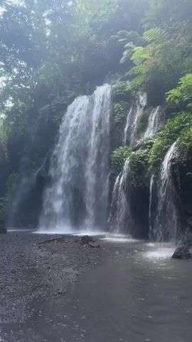 This is a vertical, eye-level shot of a waterfall surrounded by lush greenery and mist. The water cascades down a rocky cliff face, with smaller streams of water trickling down the sides. The base of the waterfall is a pool of dark water, with a rocky, dark grey shoreline. The overall impression is one of a serene and natural environment.