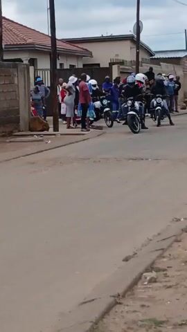Here is a description of the video. 
The video shows a group of people gathered on a street in a residential area. Three motorcycles, each with a rider wearing a helmet, drive past the group of people. The motorcycles are black and white. The riders are all wearing dark clothing. The people on the street are dressed in a variety of casual clothing. The street is paved and there are houses on either side. There is trash scattered on the side of the road.