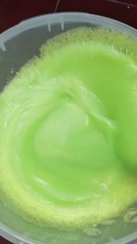 The video shows someone stirring a bucket of bright green liquid with a wooden spoon. The liquid is foamy and somewhat thick, clinging to the spoon when it is lifted. The bucket is clear plastic, and the background appears to be a dark brown surface. The stirring action is the main event taking place.