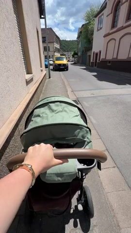 A point-of-view shot shows a person pushing a green baby stroller along a sidewalk. The person's right hand, adorned with a gold bracelet, is on the stroller's brown handlebar. The person is wearing blue jeans and white sneakers. In the background, a street with a yellow van and other cars is visible. The street slopes slightly downward, and buildings line both sides. A green, tree-covered hill is in the distance. The sky is partly cloudy. The person pushes the stroller forward, and the video ends.