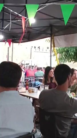 The video shows an outdoor seating area, likely a restaurant or bar, with people gathered at tables. Colorful triangular flags are strung overhead, suggesting a festive atmosphere. In the foreground, two men are seated with their backs to the camera, facing other patrons. One man wears a light-colored shirt, while the other wears a dark gray t-shirt. On the table in front of them are drinks and some food items. Further back, a woman with dark hair and glasses is visible, looking towards the camera. Another woman, also with dark hair, is seated at a table with a man, engaged in conversation. Several other individuals are seated at tables in the background, some of whom are wearing red shirts. The environment appears to be an urban or semi-urban setting, with foliage visible in the upper right corner. The lighting suggests it is daytime.