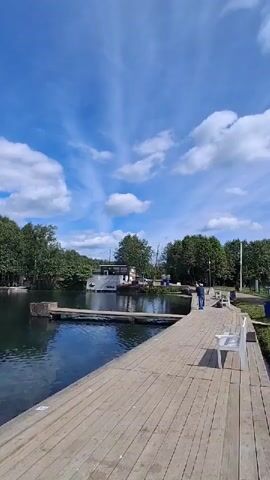 The video captures a serene daytime scene at a lake or pond in Kipen, Russia, on a bright, sunny day. A wide wooden pier extends into the dark blue-green water, occupying most of the foreground and leading towards the far bank. The sky above is a clear, vibrant blue with scattered white, wispy clouds.

On the right side of the pier, two white wooden benches are visible. A person, dressed in a blue shirt and jeans, is walking away from the camera along the pier, towards the distant end. Lush green trees line both sides of the water body, forming a verdant backdrop. Across the water, a white building with a dark roof, possibly a boathouse or recreational facility, stands near a smaller floating dock. The water appears calm, with reflections of the sky and surrounding trees. The overall atmosphere is peaceful and inviting, typical of a recreational area on a pleasant day. There is no visible text in the video.