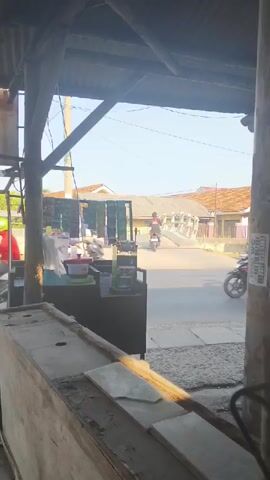 The video captures a street scene, possibly in a rural or semi-rural area, viewed from beneath a sheltered structure. The foreground is dominated by a wooden counter, partially covered by what appears to be tiling or large tiles. Behind this counter, a stall or small shop is visible, with a person in white attire serving or attending to something. Further back, the street is active with vehicular traffic. Several motorcycles are seen, some carrying passengers. A white bus with yellow accents passes by. A large red and white tanker truck is also visible. Buildings, some with pitched roofs, line the street, along with trees and utility poles. The sky is clear and blue, indicating daylight. The overall impression is a glimpse of everyday life on a street with moderate activity.