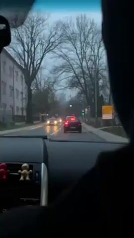 The video shows a view from inside a car looking out through the windshield. The car is stopped at a traffic light, which is green. Several other cars are ahead, also stopped at the light. The setting appears to be an urban area, with buildings and bare trees visible on either side of the road. The sky is overcast. An individual is partially visible in the foreground, likely the driver or a passenger, wearing a dark jacket or hoodie. Small decorative figures are attached to the dashboard.