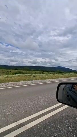 The video shows a view from the passenger side of a car driving on a two-lane road. The road has white lines marking the lanes. To the left of the road, there is a grassy field with scattered bushes and trees. In the distance, there are rolling hills covered in vegetation, and further back, larger mountains under a cloudy sky. A side mirror of the car is visible in the foreground, reflecting part of the car interior and the driver's arm. The sky is overcast with scattered clouds.