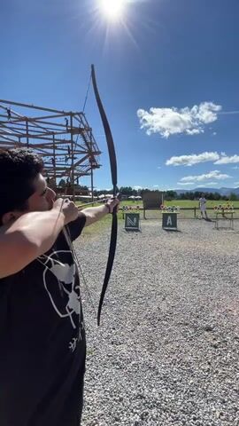 A young man in a black t-shirt with a white graphic is holding a bow and arrow, aiming at a target in the distance. He is outdoors on a sunny day with a clear blue sky and some clouds. In the background, there is a rope course structure, a gravel area, and targets set up for archery practice. Another person is visible in the distance, near the targets. The man releases the arrow and then turns to his side, looking in the direction the arrow has traveled.