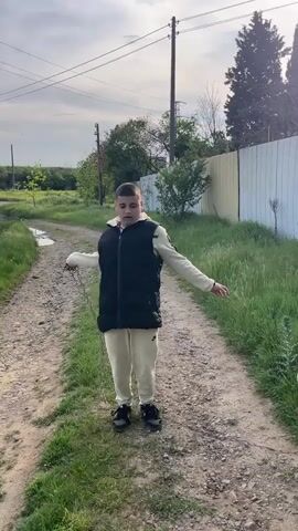 A person stands on a dirt path, flinging a chain to their side. The person is wearing a black puffy vest over a tan long-sleeved jacket, tan pants, and black shoes. To the right of the person is a white dog with its tongue hanging out. The dog then runs along the edge of the path in the grass. There is a white fence lining the path and electrical poles along the side of the path.