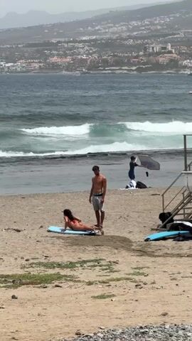 This media file captures a scene on a sandy beach with a view of the ocean and a distant town. The foreground shows two people on the beach. A woman in a red bikini is positioned on a surfboard, appearing to be in the process of practicing or learning to surf. She is on her hands and knees on the board. A man in swim trunks stands next to her, observing.

In the background, the ocean features moderate waves, suggesting conditions suitable for surfing. Further back, another person is visible near the water's edge with an umbrella. The distant shoreline is lined with buildings, indicating a populated coastal area. A lifeguard stand is also present on the beach.

The time of day appears to be daytime, and the weather seems to be overcast or hazy, with a muted quality to the light. The overall atmosphere suggests a leisurely day at the beach with some individuals engaged in water sports. The location is identified as Arona, Spain. No visible text is present in the media file.