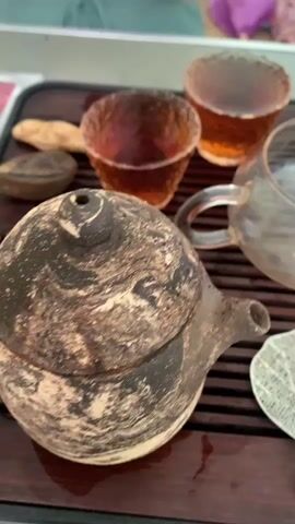 A close-up shot shows a dark, textured teapot and two amber-colored cups filled with tea. The scene is set on a dark wooden tray with grooves. A clear glass teapot with a handle is also visible, along with a white, lace-like leaf-shaped object. The background is blurred.