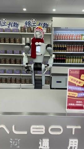The video shows a white and gray robot standing in a store in front of shelves full of products. The robot has a shiny silver head and screen displaying the words "GALBOT SERVICE" in white letters on a black background.  The robot is wearing a red scarf and a white winter hat with red and yellow trim. The shelves behind the robot are full of boxes of tea and other products. To the right of the robot is a refrigerator full of drinks. The robot is standing behind a counter. The room is well-lit and clean.