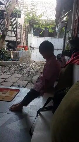 A young boy wearing a pink and white checkered shirt and dark pants sits on a white ledge. He is barefoot and has his head bowed. His right hand is brought up to his face, and he appears to be rubbing his eyes or covering them. The boy is positioned on a tiled patio area with gravel and plants visible in the background.