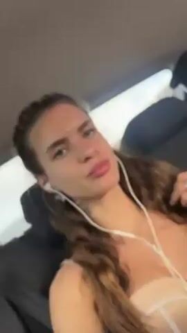 A woman with long, wavy brown hair with some strands braided back from her face sits in the back of a car. She wears white earphones and has a necklace with a key pendant. She wears a light pink top. The car seat is black, and it is raining outside. She looks at the camera with a neutral expression.