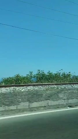 The video shows a view from inside a vehicle, likely a car, looking out the passenger window. The foreground is blurred, suggesting movement. The middle ground features a train track running horizontally across the frame, with gravel and concrete embankments. Beyond the train tracks, there is lush green vegetation and distant, hazy mountains under a clear blue sky. The camera pans slightly to the right, revealing a white concrete block with a metal box attached to it next to the train tracks. The video then shifts to an interior view of the car, showing the back of a person's head in the driver's seat, the rearview mirror, and the dashboard. The interior is also blurred, indicating continued movement.