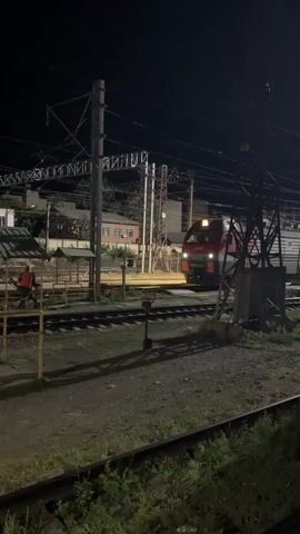 At night, a red and white train is on a railway track. A person wearing an orange vest is standing near the track on the left side of the frame, next to a covered structure. There are overhead power lines and other railway infrastructure visible. Buildings are in the background. The train's headlight is illuminated.