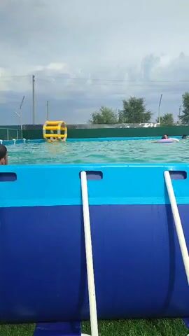 The video opens with a view of a blue and green above-ground swimming pool with several people in it, including one person on a purple inflatable. The camera then pans to the right, showing a woman in a black dress standing on a grassy area. In the background, there are more people near the pool and a large inflatable water slide with an orange and green structure. The video continues to pan right, showing more inflatable water attractions on the grass, including a large multi-colored inflatable slide and a blue inflatable pool with a canopy. Some areas of the grass appear wet, suggesting recent use of water features. The sky is cloudy.