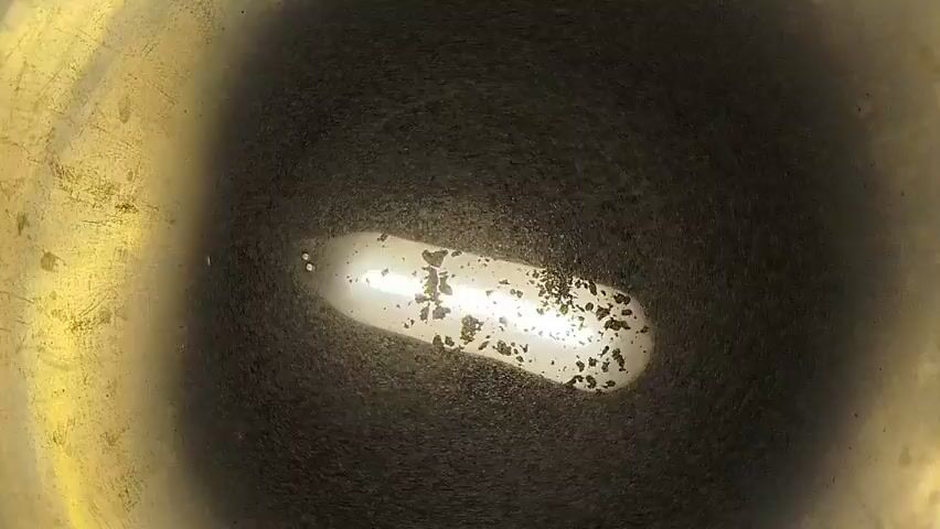 The video shows a close-up, overhead view of a dark, metallic surface, possibly the bottom of a pot or pan, with a bright, elongated light source in the center. Small, dark particles are visible on the surface, some of them clustered within the illuminated area. The edges of the surface are blurry and appear to be gold or brass colored. The camera angle remains stationary throughout the short clip.