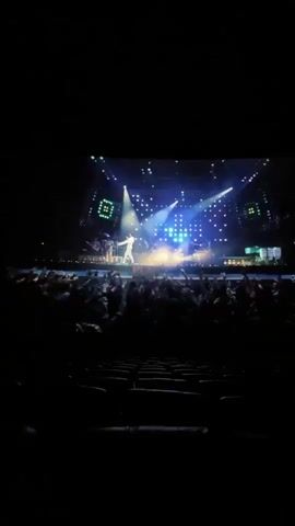 The video is shot from inside a dark movie theater. On the large screen, a recording of Michael Jackson performing is being shown. Initially, he is seen in a white outfit with striped pants, his arms outstretched to the audience. The image quickly shifts to show the screaming crowd in front of him. The dark rows of empty theater seats are visible in the foreground.