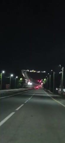 The video shows a view from a moving vehicle at night. The road ahead is illuminated by streetlights and the headlights of oncoming cars. Traffic appears to be moving slowly, with brake lights visible in the distance. The sky is dark and clear. The road has white lane markings. Trees are visible on the left side of the road, also illuminated by streetlights. The overall impression is of a typical nighttime commute or journey on a well-lit highway.