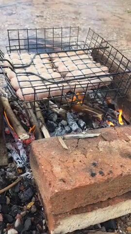 A grill basket filled with raw sausages is positioned over a campfire. The fire is burning with visible flames and embers. The background appears to be an outdoor, rocky area. There are no people or interactions visible in the video.