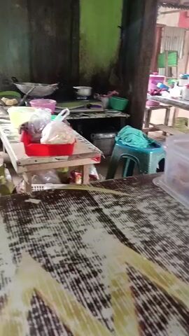 The video shows a cluttered outdoor market or food stall. In the foreground, a table is visible with a patterned surface. Beyond this table, a plastic container holding food is prominently displayed. To the left, a red tray with food is on a table. Further back, various cooking utensils, containers, and two gas cylinders are present. A turquoise stool is also visible. The background reveals shelves filled with items and a person wearing a headscarf and an orange shirt sitting on a bench. In the distance, another person is seen standing near shelves of goods. The environment appears to be a simple, functional setup, possibly for selling food.