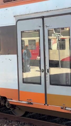 The video shows a white and orange train with peeling paint. The doors of the train have large windows that reflect the image of another train. Two people are reflected in the windows. One person is wearing a blue jacket and the other is wearing a white hat. The train is on a track.