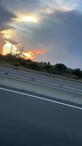 The video shows a view from a vehicle traveling on a highway. The sky is cloudy with the sun breaking through, casting rays of light and creating an orange and pink hue. Several vehicles are visible on the highway, including a white car in the left lane and a white truck in the right lane. The camera appears to be moving, causing motion blur. The environment includes a highway with white lane markings and a shoulder, with some trees and hills in the background.