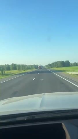 A vehicle is driving on a highway under a clear blue sky. The road is paved and has white lane markings. On either side of the road, there are fields of green grass and trees. There are other vehicles on the road, including trucks. The camera appears to be mounted inside the vehicle, looking forward.