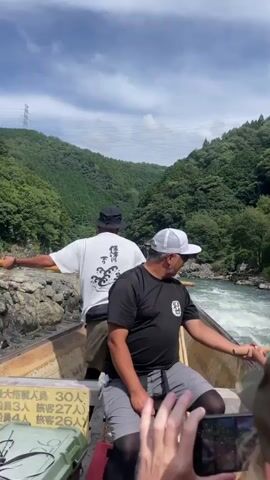 This video captures a scenic boat ride down a river. In the foreground, we see the backs of two passengers. A man in a white cap and black t-shirt is steering the boat with a long wooden oar. Behind him, another man in a black hat and white t-shirt also holds an oar. In front of them, other passengers are visible, with one woman in a green cap holding a phone up to record. The boat is wooden and appears to be carrying multiple people. The river is wide and choppy with whitewater, suggesting a fast current. The surrounding environment is lush and green, with steep, tree-covered hills on either side of the river, and a cloudy blue sky above. There are some rock formations visible along the riverbanks, and a power line pylon can be seen in the distance on the left side of the frame. A sign with Japanese characters is attached to the side of the boat. The camera is positioned from the perspective of someone within the boat, looking forward. The overall impression is one of an adventurous and picturesque journey.