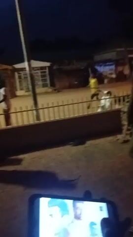 The video is taken at night and appears to be filmed from a low angle, possibly from a phone. In the foreground, a person's hand is holding a smartphone, displaying an image that is not clear due to the low resolution and lighting. The background shows an outdoor scene with several individuals. In the center of the frame, a person wearing a yellow shirt and dark pants is facing away from the camera and appears to be in motion, possibly dancing or performing an action. To the left, two individuals are partially visible; one is wearing a light-colored garment and the other is wearing a light-colored shirt and dark pants. Further to the right, there is a white car parked behind a fence. The environment includes a fence, buildings with open structures, and a pole with a light. The ground is paved. The overall scene is dimly lit, suggesting it is nighttime.