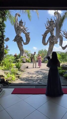 The video shows a person taking pictures of a couple in an outdoor garden area. There are two tall sculptures of figures in dancing poses on either side of a stone path. The couple is standing on the path holding hands and posing for the photo. The woman is wearing a light pink dress, and the man is wearing a green shirt and tan pants. The person taking the picture is standing on a red rug inside of a building, looking out at the garden. The sky is blue and clear. There are palm trees and other plants in the background.
