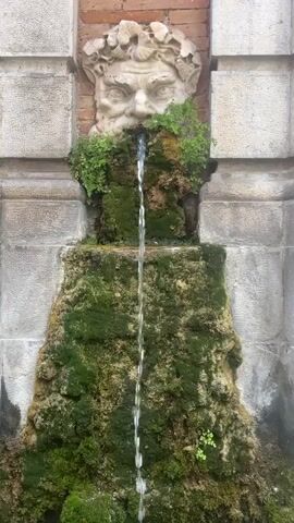 A water fountain featuring a sculpted head with a beard and leaves around its head is depicted. Water flows from the mouth of the head down a moss-covered rock structure. The fountain is set against a wall made of brick and stone blocks. The video is shot from a stationary position directly in front of the fountain.