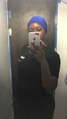 A person stands in a public restroom, taking a selfie in the mirror with their smartphone. The person has dark skin and is wearing a blue head covering and a black T-shirt with a white logo on the left side. The phone held by the person is grey with an Apple logo visible. The restroom has white and grey tiled walls. The mirror reflects a door with a blue frame and the interior of the restroom stalls. The person's face becomes visible as they lower the phone. The person appears to look at themselves in the mirror before returning to looking at the phone.