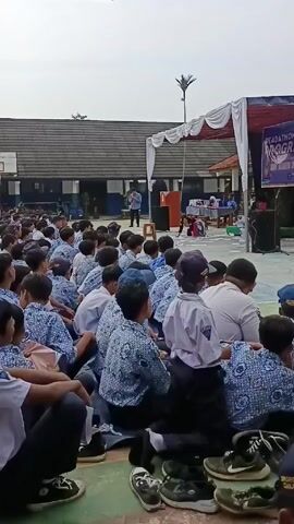 A medium shot shows a crowd of children sitting on the ground and listening to a man speaking on a stage. The children are wearing school uniforms of blue and white. Many of them are sitting with their knees drawn up, while others have their legs extended.  The stage is decorated with white and red fabric. There is a banner on the right side of the stage. A man stands at the lectern, speaking to the crowd. Behind the crowd is a school building with a basketball court. The sky is overcast. There are many shoes scattered in the foreground, apparently left by the students.