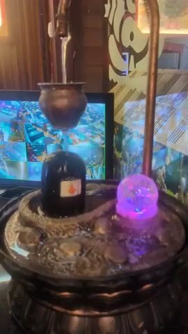 A tabletop fountain features a black lingam topped with a small copper pot from which water streams down. The lingam is encircled by a coiled snake. To the right of the lingam, a translucent sphere glows with purple light. The fountain sits in a dark, ornate base. In the background, a computer monitor displays a grid of security camera feeds. The environment appears to be indoors, with wooden paneling and a decorative wallpaper visible.