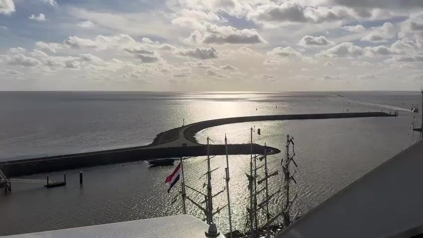 The video shows a wide shot of a harbor with a curved breakwater. The sun is shining brightly on the water, creating a glare. A large sailboat with multiple masts is docked in the foreground, and a Dutch flag is visible on the boat. A road is visible on top of the breakwater. In the distance, a long pier extends into the sea, with a faint line of lights visible along it. The sky is partly cloudy, with the sun breaking through the clouds. The overall scene is calm and serene, with a sense of vastness.
