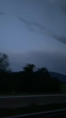 The video shows a nighttime scene from a vehicle. A dark silhouette of trees and a distant mountain range is visible against a dusky sky. A bright light, possibly a star or planet, is visible in the sky. The road is visible in the foreground. The video is blurry, suggesting movement.
