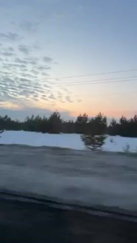 The video depicts a view from a moving vehicle driving alongside a snowy field. A line of trees is visible in the distance. The sky is blue with some clouds and the sun is setting, casting an orange and pink glow on the horizon. There are power lines running above the scene. The road is visible in the foreground, showing streaks of dirt or slush.