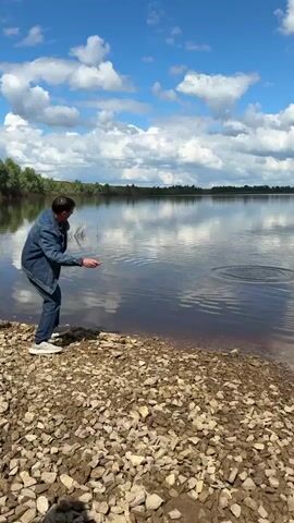 A man throws something into a lake. The man is wearing a denim jacket, jeans, and sneakers. He stands on a rocky beach. A child walks up next to him. The lake is calm and reflects the blue sky and clouds. There are trees in the background. The man is looking at the lake. He appears to have thrown something into the lake, and it made ripples.