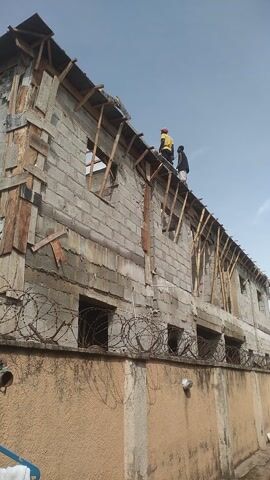 The video shows a tall, unfinished building under construction. The exterior walls are made of concrete blocks. Two construction workers are visible near the top of the building, on the roof structure, which is supported by wooden beams. Barbed wire is strung along the upper floors of the building, suggesting a security measure or perhaps part of the construction. The sky is clear and blue. The overall scene depicts an active construction site.