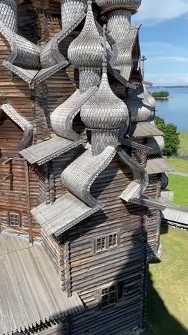 A close-up, aerial shot pans across the ornate wooden architecture of a historical church, likely in Northern Russia, given the style. The camera focuses on the intricate details of the structure, showcasing its many domes, onion-shaped roofs, and layered wooden shingles, all weathered and aged. The detailed woodwork, characteristic of traditional Russian wooden architecture, is the primary subject, with the natural elements like the blue sky, clouds, and a body of water with distant islands forming the background. The shot emphasizes the scale and complexity of the building, hinting at its cultural and historical significance.