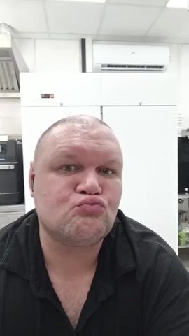 A man with a bald head and a black shirt is making faces at the camera. He has his eyes closed and his lips pursed in the first frame. He then opens his eyes wide and his lips remain pursed in the subsequent frames. He appears to be in an indoor environment with kitchen appliances in the background.