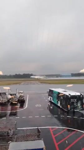 An airplane, white with green tail markings, is captured taking off from a wet runway at an airport on an overcast day. The aircraft is shown ascending with visible exhaust or water vapor trailing from its engines. In the foreground and midground, various airport ground support equipment, including baggage carts and a large airport bus, are parked on the dark, wet tarmac, which has red and white markings. The scene is viewed from an indoor location, evidenced by reflections on the window through which the video is recorded. Trees are visible in the distant background beyond the runway.