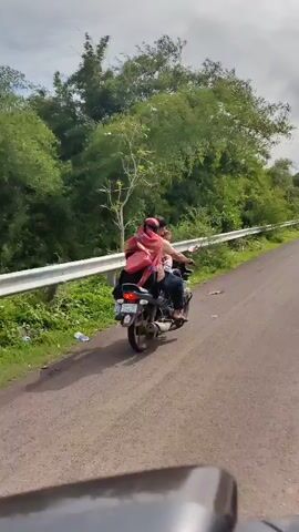 A man and two children are riding on a motorcycle on a paved road. The man is driving, and one child is sitting in front of him. The other child is sitting behind the man. All three individuals are looking at the camera. The motorcycle is moving forward, and the background consists of a lush green environment with trees and foliage. The road appears to be in a rural or natural setting.