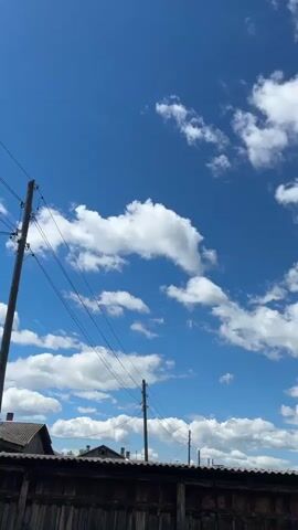 The video shows a view of a bright blue sky filled with fluffy white clouds. In the foreground, there is a wooden fence and the roof of a building. Power lines and utility poles are visible against the sky. The camera is looking upwards, with the sky dominating the frame. The overall impression is of a clear, sunny day.
