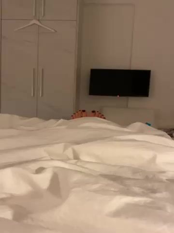 A camera is pointed at a bed covered in white sheets. Two feet, with black painted toenails, are sticking out from underneath the covers towards the top of the bed.  Behind the head of the bed is a pillow, a wall-mounted television, and a closet. A hanger hangs inside the closet.